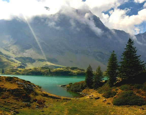 Idyllische Berglandschaft mit einem t&uuml;rkisfarbenen See im Vordergrund, umgeben von gr&uuml;ner Vegetation und Nadelb&auml;umen. Im Hintergrund ragen wolkenverhangene Berge empor, durch die Sonnenstrahlen brechen.