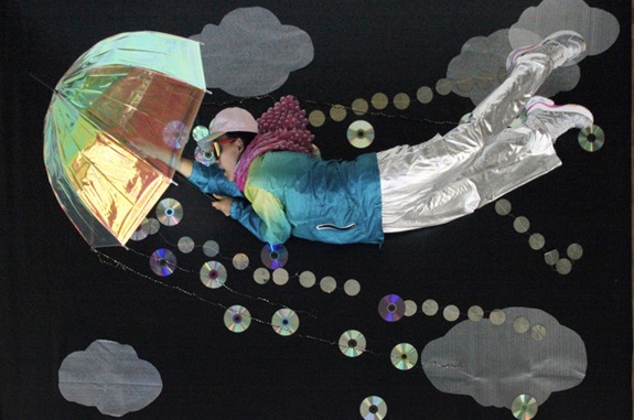 Person in schillerndem Outfit fliegt mit transparentem Schirm durch eine dunkle Szene, umgeben von Wolken und Ketten aus CDs.