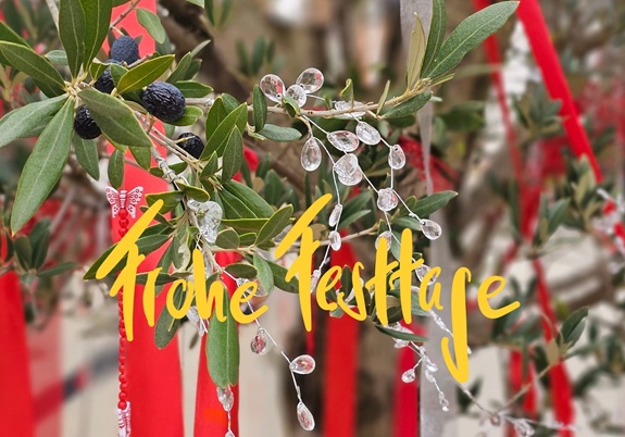 Ein Olivenzweig mit schwarzen Fr&uuml;chten und Kristall-Dekoration, im Hintergrund rote B&auml;nder. Gelber Schriftzug: &bdquo;Frohe Festtage&ldquo;.
