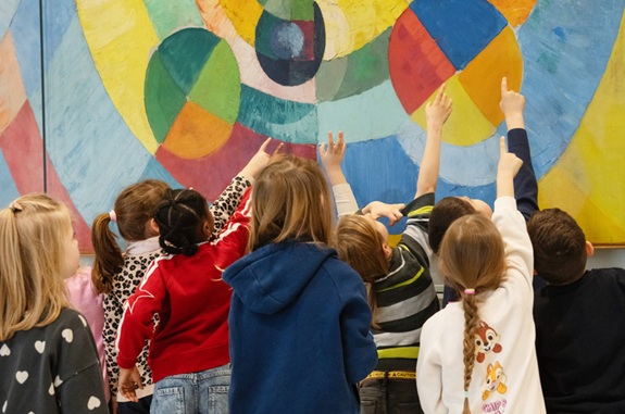 Kindergruppe betrachtet ein gro&szlig;es, buntes abstraktes Gem&auml;lde und zeigt mit erhobenen Armen darauf.
