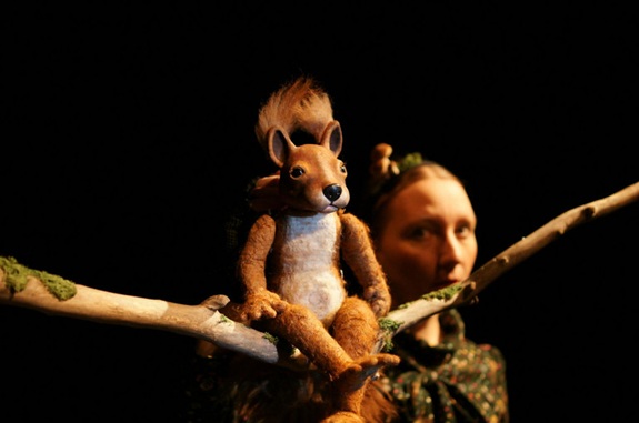Detailreiche Handpuppe eines Eichh&ouml;rnchens, die auf einem Ast sitzt. Im Hintergrund ist die Puppenspielerin oder der Puppenspieler sichtbar.