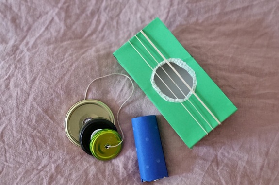 Selbstgebastelte Musikinstrumente aus Alltagsmaterialien: eine gr&uuml;ne Schachtel mit Gummib&auml;ndern (&auml;hnlich einer kleinen Gitarre) und ein Objekt aus Deckeln und Schnur.