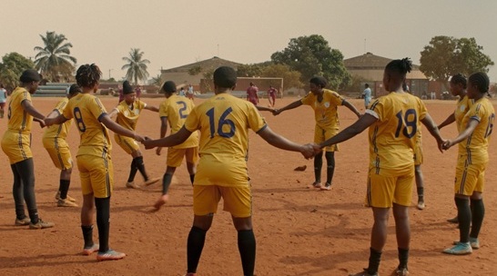 Afrikanische Fussballerinnen in gelben Trikots halten sich in einem Kreis an den H&auml;nden.  Sie stehen auf roter Erde.