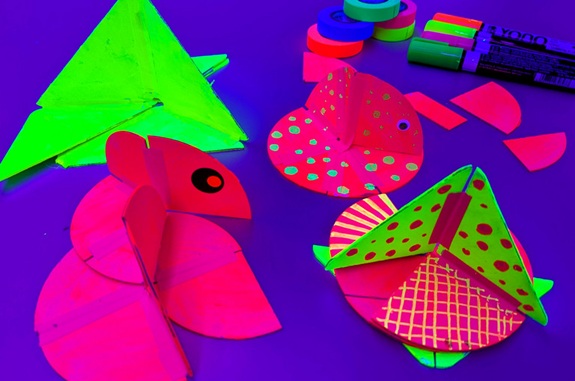 Bunte Papierbasteleien in Neonfarben (Fische und Formen) mit Klebeb&auml;ndern und Markern auf violettem Untergrund.