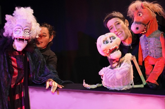 Eine Puppenspielszene mit bunten Figuren, darunter ein Skelett in weissem Tuch und eine Pferdepuppe in rotem Outfit.