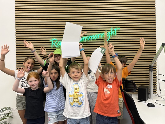 Eine Gruppe von Kindern in einem Raum mit Holzlamellenwand und gr&uuml;nem Neon-Schriftzug. Sie halten Bl&auml;tter hoch und strecken jubelnd die Arme in die Luft.
