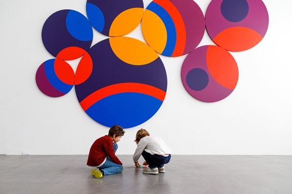 Zwei Kinder, die auf dem Boden sitzen und etwas betrachten. Im Hintergrund h&auml;ngt eine gro&szlig;e Wandinstallation aus bunten, &uuml;berlappenden Kreisen in Blau, Rot, Orange und Violett. 