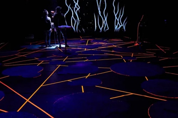 Dunkler Raum mit beleuchtetem Boden (orange Linien und Kreise), zwei Personen im Hintergrund, abstrakte Lichtmuster an der Wand.