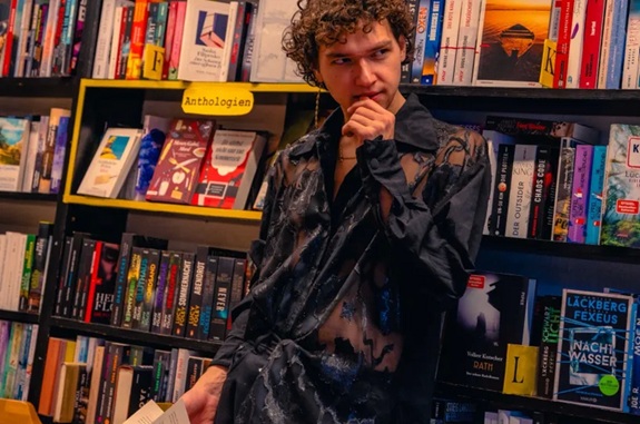 Eine Person steht in einem Buchladen vor vollen Regalen und h&auml;lt ein ge&ouml;ffnetes Buch in der Hand. Im Hintergrund sind viele B&uuml;cher sichtbar, darunter ein Regal mit der gelben Beschriftung &bdquo;Anthologien&ldquo;.