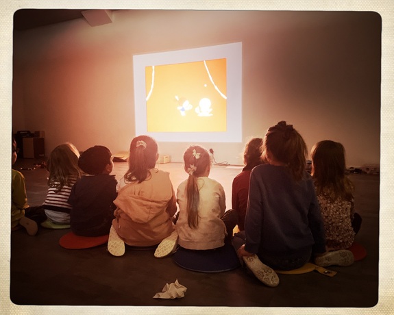 Eine Gruppe von Kindern sitzt auf bunten Sitzkissen in einem abgedunkelten Raum und schauen gemeinsam eine Projektion auf einer wei&szlig;en Wand an. Die Projektion zeigt eine einfache, orangefarbene Zeichentrickszene.
