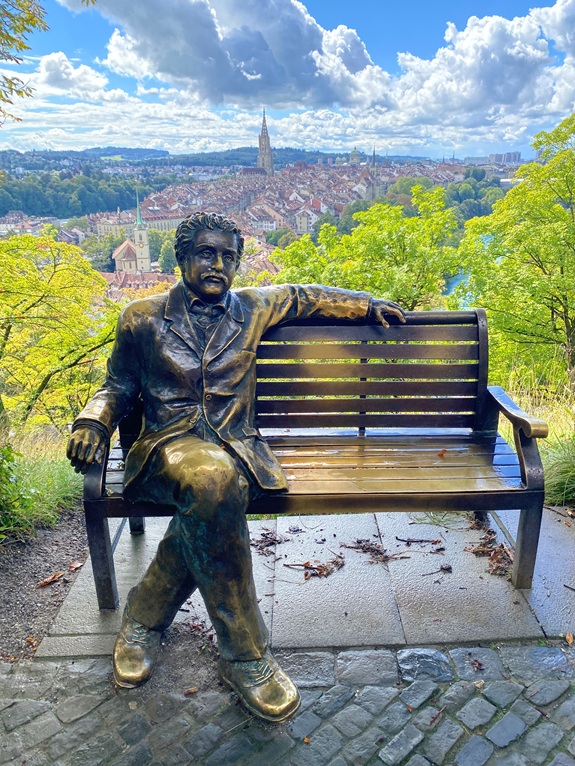 Eine bronzene Statue (Einstein) sitzt entspannt auf einer Holzbank, dahinter eine Stadtansicht (Bern) mit roten D&auml;chern, Kirchturm und gr&uuml;nen B&auml;umen unter blauem Himmel mit Wolken. 