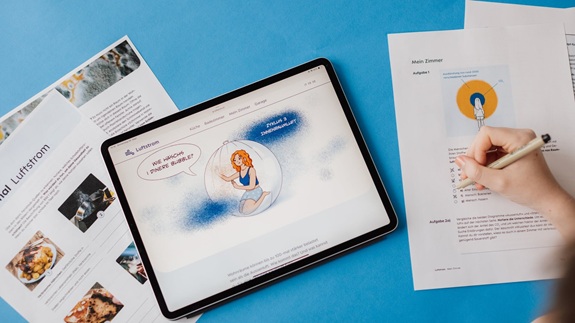 Ein Tablet mit einer Illustration und Text liegt auf einem blauen Untergrund, daneben ausgedruckte Seiten und eine Hand, die auf einem Blatt schreibt.