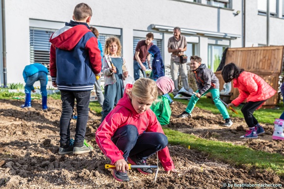Kinder arbeiten gemeinsam in einem Gartenbeet vor einem Geb&auml;ude, einige knien und benutzen kleine Werkzeuge, andere stehen und helfen. (Foto: Bea Habermacher)