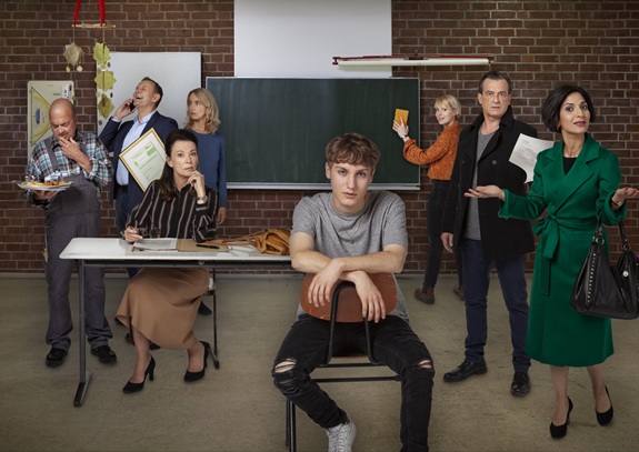 Eine Gruppe von Personen steht und sitzt in einem Klassenraum; einige sprechen, gestikulieren oder halten Unterlagen, w&auml;hrend eine Person l&auml;ssig auf einem Stuhl im Vordergrund sitzt.