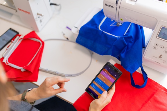 Eine Person steuert eine N&auml;hmaschine mit Stoff und Stickrahmen &uuml;ber ein Smartphone.