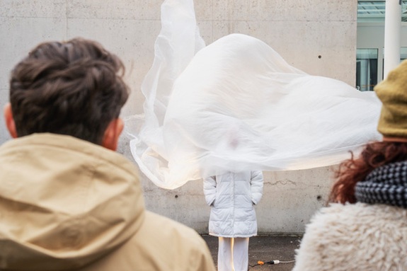 Eine Performance im &ouml;ffentlichen Raum: Eine Person steht vor einer Betonwand, umh&uuml;llt von einem grossen, weissen, im Wind wehenden Stoff, beobachtet von Zuschauenden.
