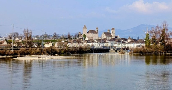 Blick &uuml;ber einen ruhigen See auf eine historische Altstadt mit Kirche und Bergen im Hintergrund.
