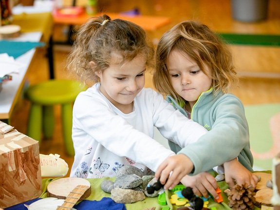 Zwei Kinder spielen gemeinsam an einem Tisch mit Naturmaterialien und Bastelobjekten in einem Innenraum.