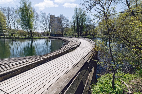 Geschwungener Holzsteg entlang eines Teichs in einem gr&uuml;nen Park mit B&auml;umen und Geb&auml;uden im Hintergrund.