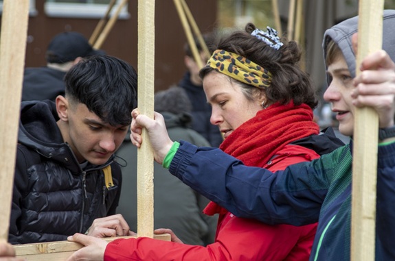 Mehrere Personen halten gemeinsam Holzst&auml;be bei einer Veranstaltung im Freien.
