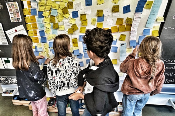 Vier Kinder stehen vor einer Wand voller bunter Post‑its und ordnen sie gemeinsam.