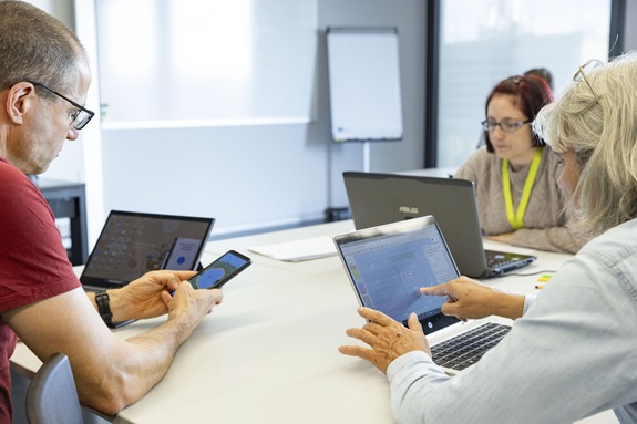 Drei Erwachsene sitzen an einem Tisch und arbeiten gleichzeitig mit Laptop und Smartphone.