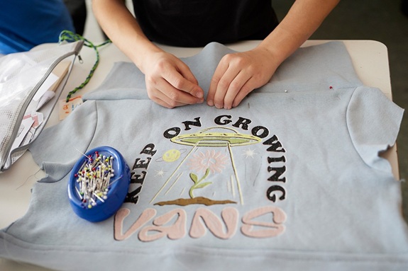 H&auml;nde stecken Stecknadeln in ein T‑Shirt mit Aufdruck auf einem Tisch.