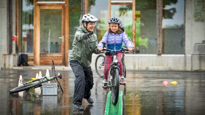 Eine erwachsene Person unterst&uuml;tzt ein Kind dabei, auf einem Fahrrad &uuml;ber eine schmale, gr&uuml;ne Rampe zu fahren &ndash; offenbar bei einem Fahrtraining im Freien auf nasser Fl&auml;che.