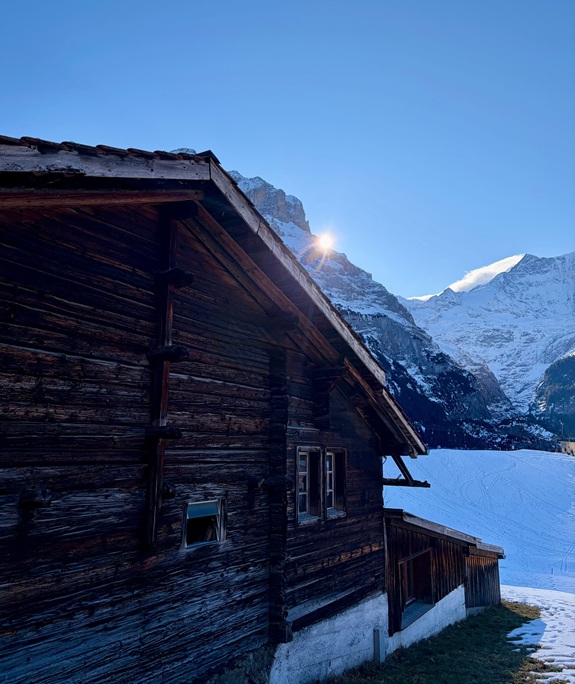 Eine Holzbergh&uuml;tte steht im Vordergrund, dahinter erheben sich schneebedeckte Berge unter klarem blauem Himmel, mit Sonnenschein am Gipfelrand.