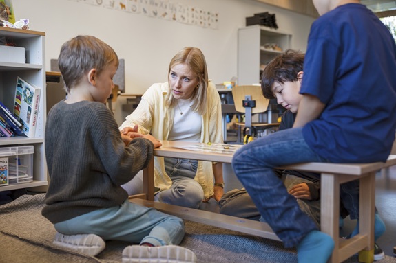Eine Lehrerin sitzt mit drei Kindern in einem Klassenzimmer am kleinen Tisch und erkl&auml;rt ihnen etwas mit Lernmaterial. Die Kinder h&ouml;ren aufmerksam zu und arbeiten gemeinsam.