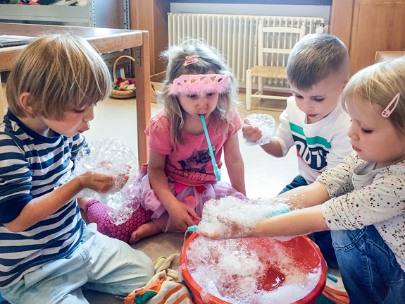 Vier Kinder sitzen am Boden und spielen mit Schaum, den sie aus einem Becken vor ihnen haben.