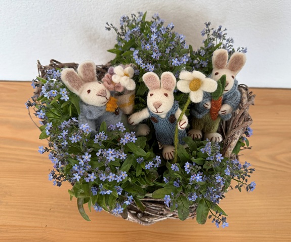 Drei kleine Filzhasen mit Blumen sitzen in einem Nest aus gr&uuml;nen Pflanzen und blauen Bl&uuml;ten. 