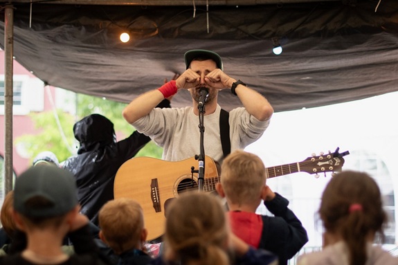 Person mit Gitarre auf einer kleinen B&uuml;hne macht Gesten vor einem Publikum aus Kindern.