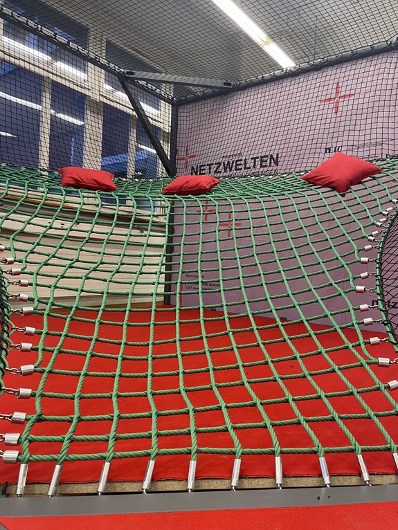 Gr&uuml;nes Kletternetz mit roten Polstern in einem Innenraum, beschriftet mit &laquo;NETZWELTEN&raquo;.