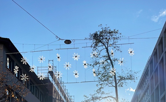 Lichtergirlanden in Bl&uuml;tenform h&auml;ngen &uuml;ber einer Strasse zwischen Geb&auml;uden vor blauem Himmel und einem Baum.