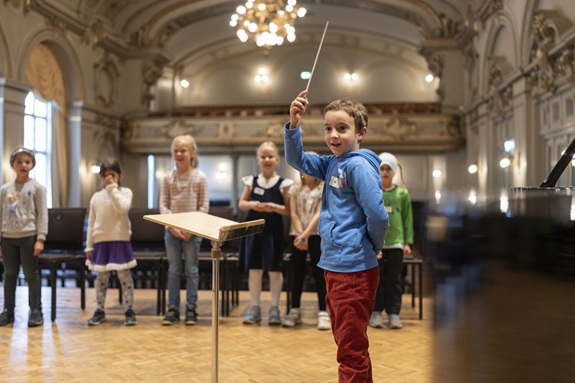 Kind dirigiert in einem Konzertsaal eine Gruppe von Kindern auf der B&uuml;hne.