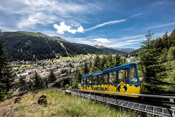Zahnradbahn (Schatzalpbahn) f&auml;hrt durch bewaldeten Hang &uuml;ber einem Tal mit Bergdorf und Alpen im Hintergrund.