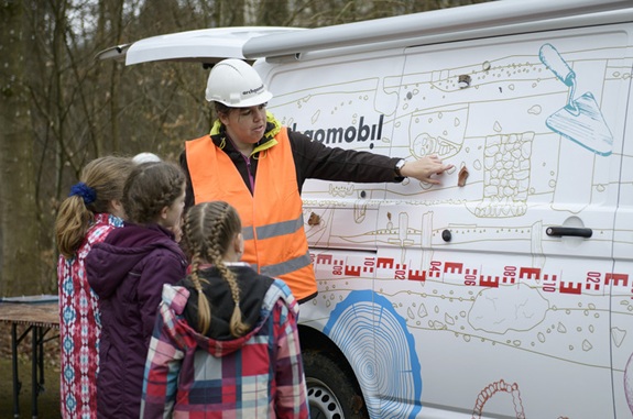 Eine Person mit Sicherheitsweste und Helm erkl&auml;rt einer Gruppe von Kindern etwas an der bebilderten Au&szlig;enwand eines Fahrzeugs.