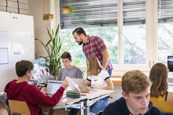 Lehrperson unterst&uuml;tzt Sch&uuml;lerinnen und Sch&uuml;ler beim Arbeiten mit Laptops im Klassenzimmer.