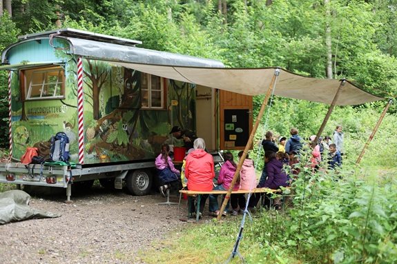 Kinder sitzen vor einem Bauwagen im Wald und h&ouml;ren einer Outdoor‑Lektion zu.