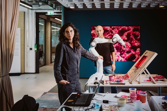 Eine Person arbeitet in einem modernen Atelier mit einem Roboterarm an einem kreativen Kunstprojekt, umgeben von Farben und technischem Equipment.