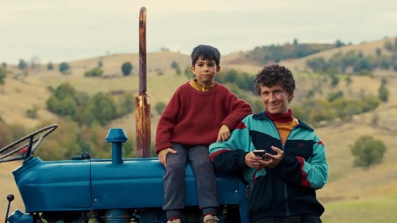 Zwei Kinder sitzen auf einem Traktor in einer l&auml;ndlichen Landschaft mit H&uuml;geln im Hintergrund.