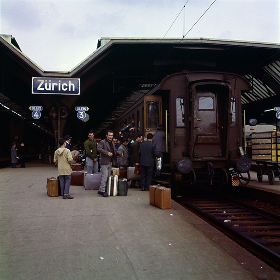 Historische Szene am Z&uuml;rcher Bahnhof mit einem Zug am Perron und Reisenden mit Koffern beim Einsteigen.