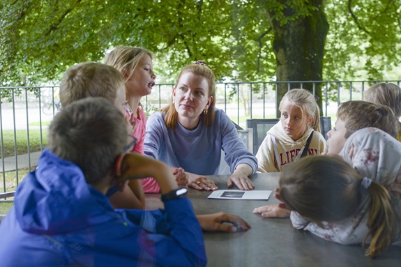 Gruppe von Kindern sitzt mit einer Erwachsenen an einem Tisch im Freien und bespricht etwas gemeinsam.