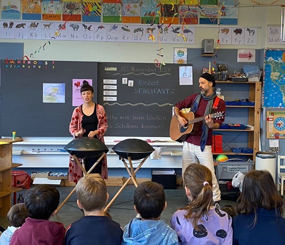 Musikauff&uuml;hrung im Klassenzimmer: Zwei Personen spielen Instrumente vor sitzenden Kindern.