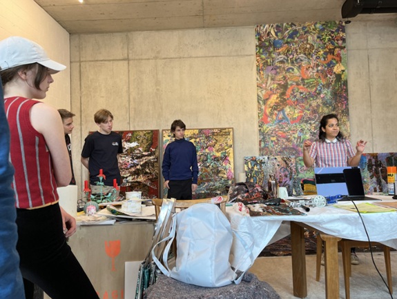 In einem Atelier stehen mehrere Personen zwischen Tischen voller Farben und Materialien, w&auml;hrend im Hintergrund farbenfrohe, abstrakte Gem&auml;lde an der Wand h&auml;ngen. Eine Person vorne rechts scheint etwas zu erkl&auml;ren oder vorzustellen.