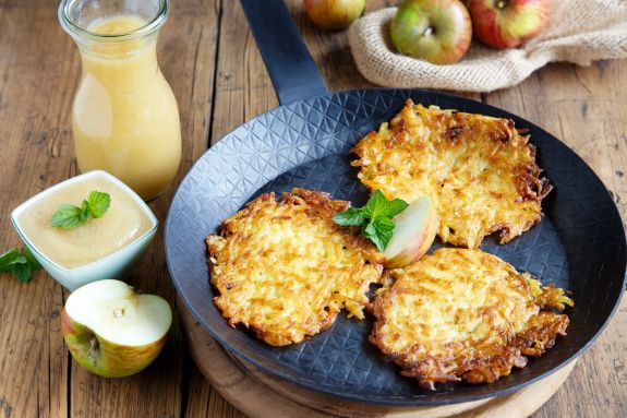 R&ouml;sti auf einem Teller, daneben Apfelmus, Apfelsaft und ein halber Apfel.