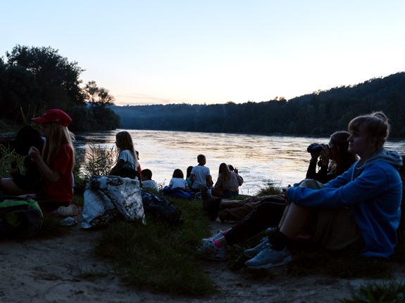 Jugendliche sitzen am Flussufer und schauen bei D&auml;mmerlicht auf das Wasser.