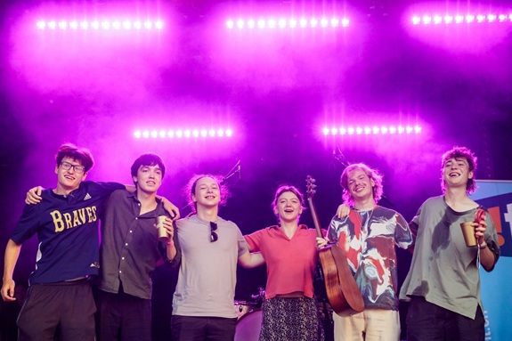 Eine Gruppe von sechs Personen steht eng beisammen auf einer B&uuml;hne, umgeben von violettem Scheinwerferlicht. Einige halten Getr&auml;nke, eine Person tr&auml;gt eine Gitarre. Die Stimmung wirkt fr&ouml;hlich und gemeinschaftlich.