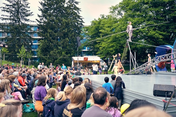 Open-Air-Auff&uuml;hrung mit Seiltanz und Akrobatik auf einer B&uuml;hne, beobachtet von einem gro&szlig;en Publikum im Park.
